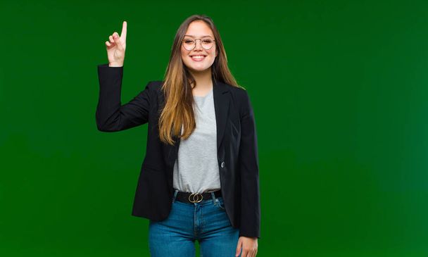 jonge mooie vrouw glimlachen vrolijk en gelukkig, wijzen naar boven met een hand om ruimte te kopiëren tegen groene achtergrond - Foto, afbeelding