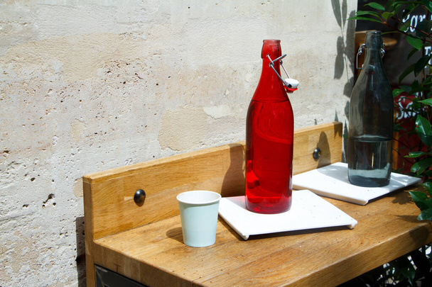 Close-up of a red water bottle and glass on a restaurant table against a light stone wall - 写真・画像