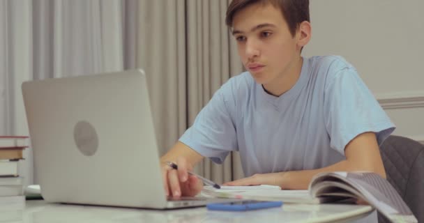 Teen boy doing school lessons at home. Student writes the homework. Child learns. Boy using laptop. Student writes with a pen in a notebook. - Кадры, видео