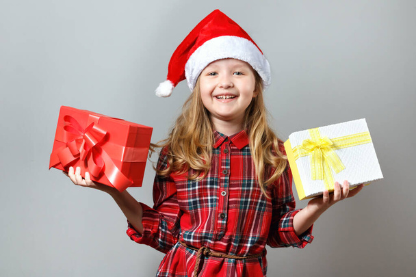 Linda niña encantadora en un sombrero de santa y vestido rojo sobre un fondo gris. El niño sostiene dos cajas con regalos en sus manos. Navidad y año nuevo concepto
 - Foto, imagen