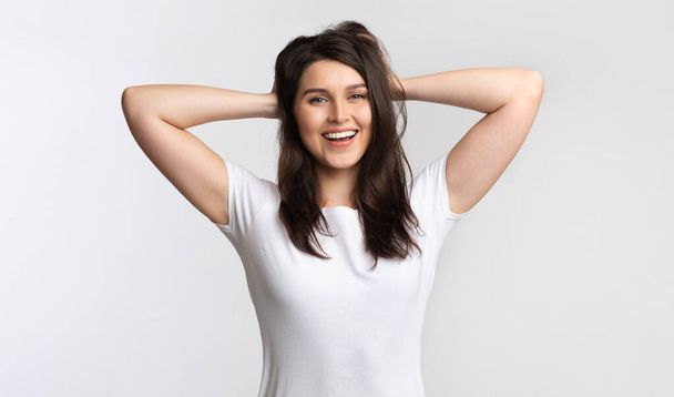 Chica positiva tocando el cabello de pie posando sobre fondo blanco, estudio
 - Foto, Imagen