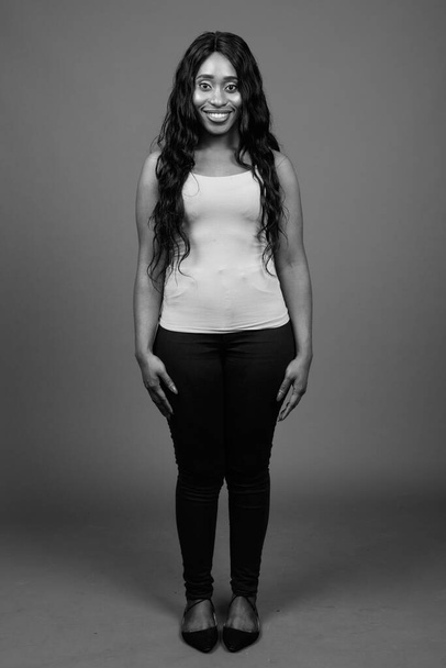 Studio shot της νεαρής όμορφης Αφρικανικής γυναίκας με μακριά σγουρά μαλλιά σε γκρι φόντο σε μαύρο και άσπρο - Φωτογραφία, εικόνα