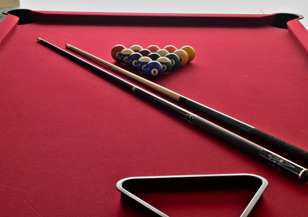 Colored and numbered billiard balls, black ball rack, and two pool cues on a red felt pool table. - 写真・画像