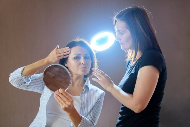 sprechende und lächelnde Kosmetikerin und reife Patientin mit Spiegel im Schönheitssalon - Foto, Bild