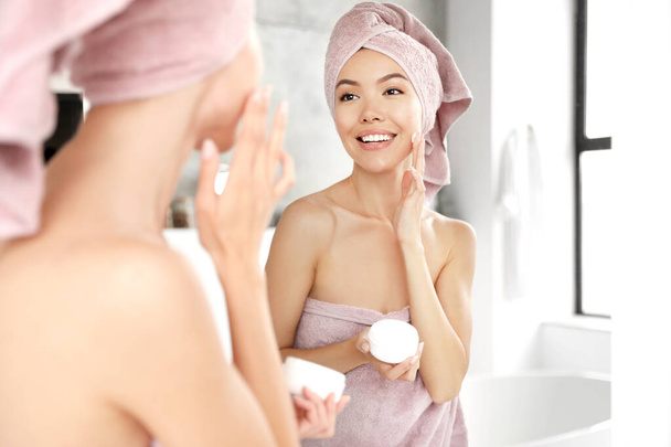 Young woman with towels applying facial cream near mirror in bathroom - Fotó, kép