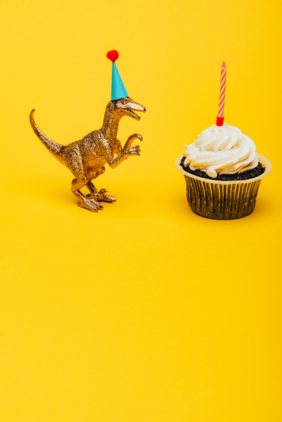 Dinosaurio de juguete en gorra de fiesta junto a magdalena con vela sobre fondo amarillo
 - Foto, Imagen
