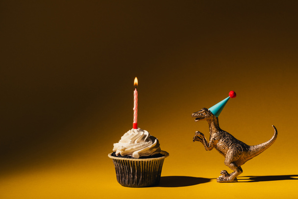 Dinosaurio de juguete en gorra de fiesta junto a magdalena con vela encendida sobre fondo marrón
 - Foto, imagen