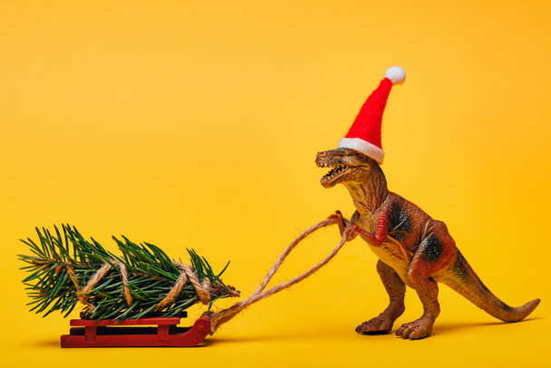 Speelgoed dinosaurus in santa hoed met dennen op slee op gele achtergrond - Foto, afbeelding