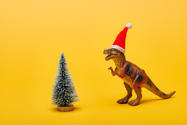 Toy dinosaur in santa hat beside christmas tree on yellow background - Photo, Image