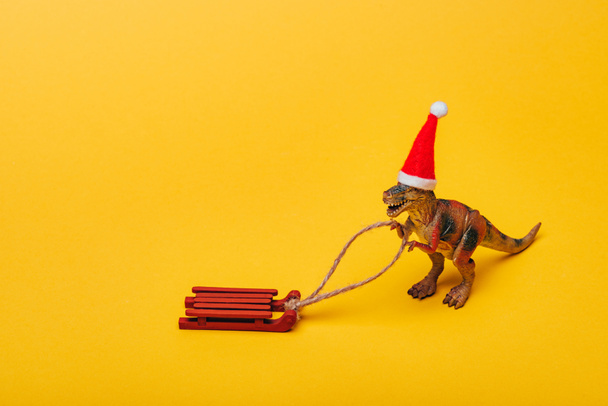 Toy dinosaur in santa hat with sleigh on yellow background - Photo, Image