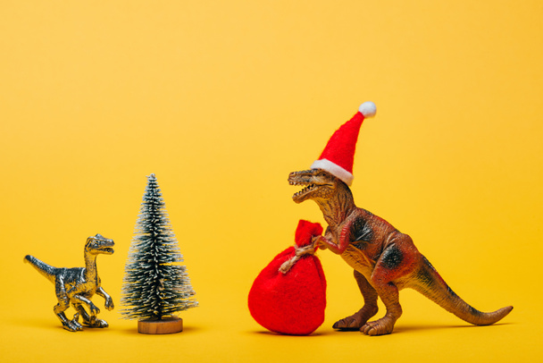 Toy dinosaurs with santa hat and sack beside pine on yellow background - Photo, Image