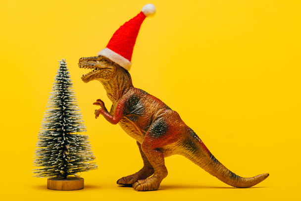 Toy dinosaur in santa hat beside christmas tree on yellow background - Foto, Imagem