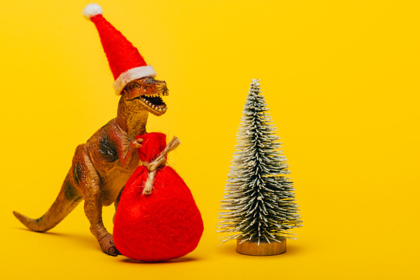 Toy dinosaur in santa hat with sack beside christmas tree on yellow background - Фото, зображення