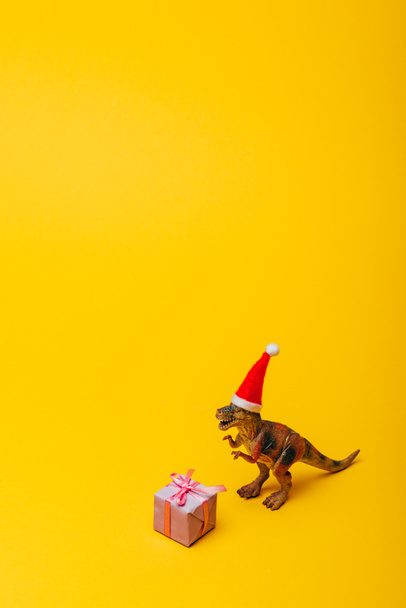 Toy dinosaur in santa hat with gift on yellow background - Фото, изображение