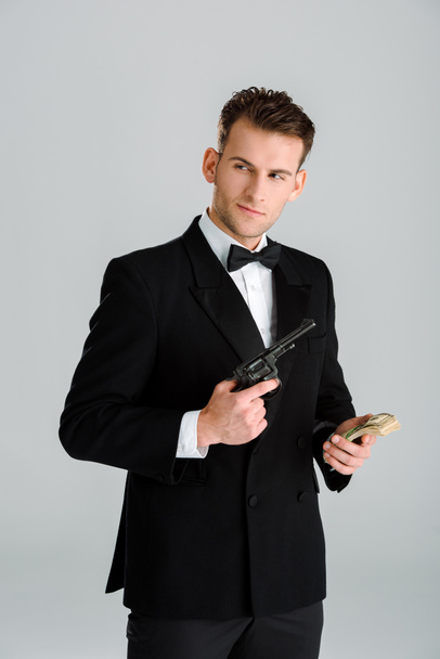 hombre guapo sosteniendo pistola y billetes de dólar aislados en gris
  - Foto, Imagen