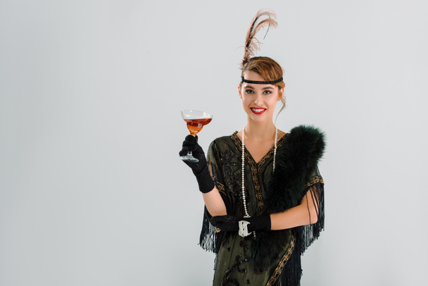 cheerful aristocratic woman holding glass with alcohol drink isolated on grey  - Photo, Image