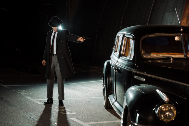 komea gangsteri hattu ja takki pitämällä ase lähellä retro auto
  - Valokuva, kuva