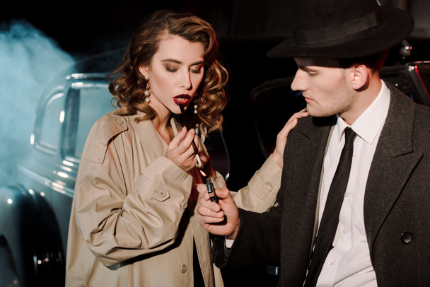 όμορφος γκάνγκστερ στο καπέλο κρατώντας αναπτήρα κοντά ελκυστική γυναίκα καπνίζει κοντά στο αυτοκίνητο σε μαύρο με καπνό  - Φωτογραφία, εικόνα