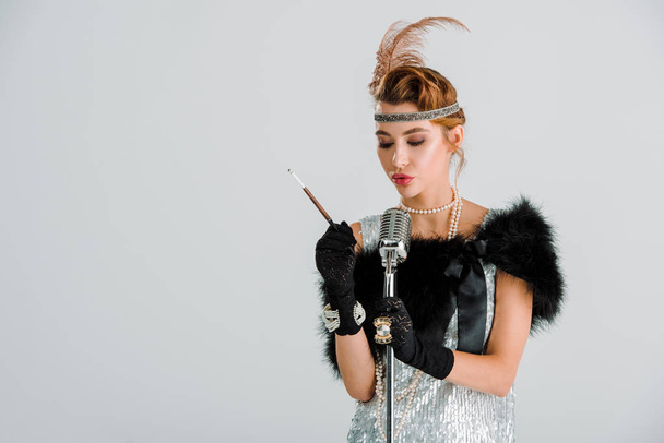 attractive woman touching retro microphone and holding cigarette holder isolated on grey  - Фото, зображення