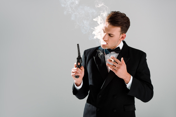 опасный человек в костюме с галстуком-бабочкой, держащий пистолет, пока курит сигару, изолированную на сером
  - Фото, изображение