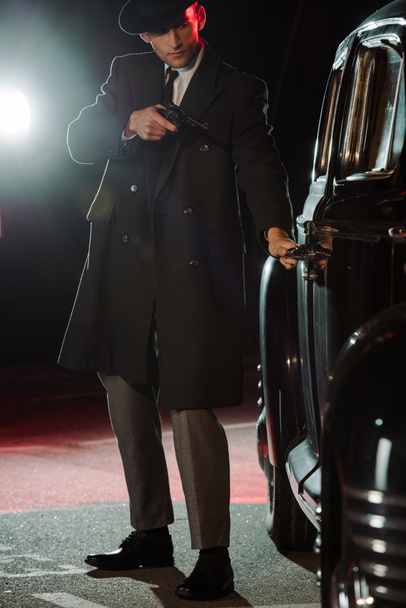 stylish gangster in hat and coat holding gun near retro black car  - Zdjęcie, obraz