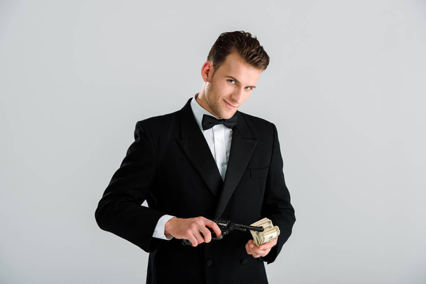 šťastný zločinec držící zbraň a dolarové bankovky izolované na šedi  - Fotografie, Obrázek