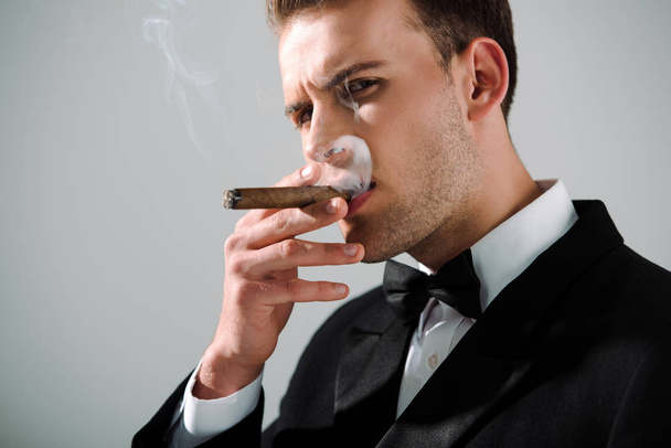 όμορφος και πλούσιος άντρας καπνίζει πούρο απομονωμένος στο γκρι  - Φωτογραφία, εικόνα