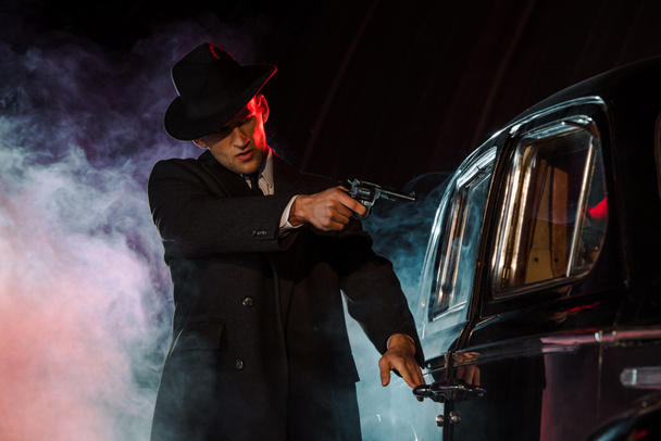 stylish gangster holding gun near retro car on black with smoke  - Fotoğraf, Görsel