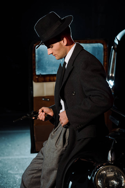 side view of handsome man in hat holding gun near vintage car  - Photo, Image
