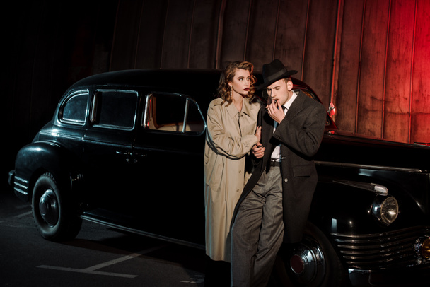όμορφος άντρας με καπέλο καπνίζει πούρο κοντά σε ελκυστική γυναίκα και vintage αυτοκίνητο  - Φωτογραφία, εικόνα