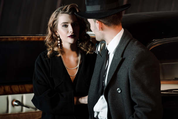 selective focus of attractive woman standing with gangster in hat near retro car  - Фото, изображение