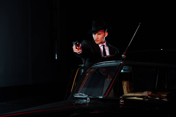 コートを着た武装した男とレトロな車の近くに銃を持った帽子  - 写真・画像