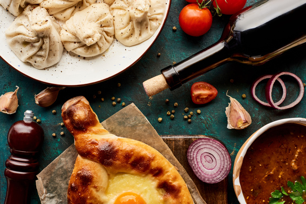 top view of delicious khinkali, Adjarian khachapuri, kharcho and bottle of wine on green background - Photo, Image