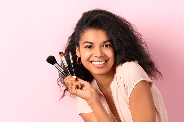 Mooie Afro-Amerikaanse vrouw met make-up borstels op kleur achtergrond - Foto, afbeelding