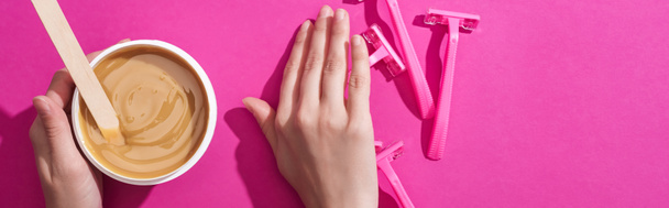 cropped view of woman moving away disposable razors and taking depilation wax in cup with stick on pink background, panoramic shot - Foto, Imagen
