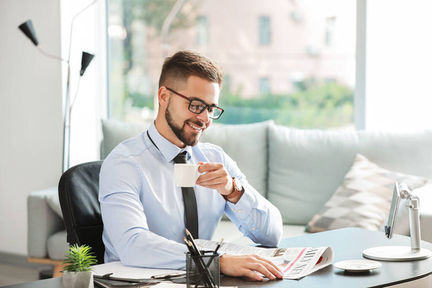 Красивый бизнесмен пьет кофе во время чтения газеты в офисе
 - Фото, изображение