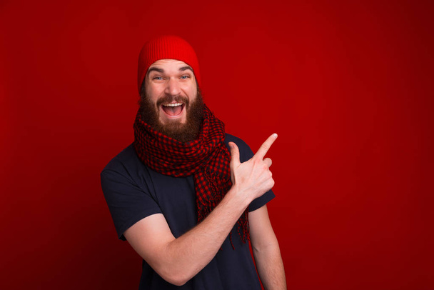 Ευτυχισμένος τύπος που φοράει κόκκινο καπέλο και μαντήλι, δείχνοντας αλλού ενώ κοιτάζει - Φωτογραφία, εικόνα