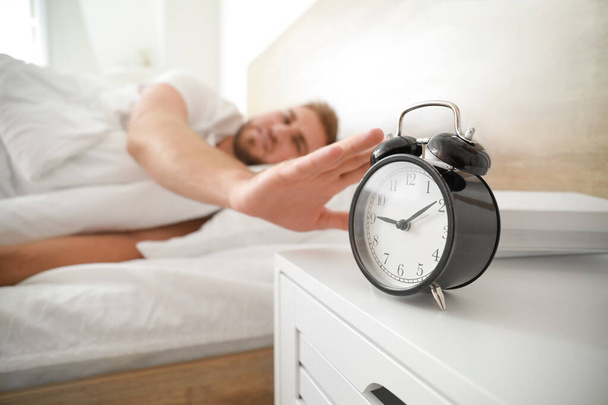 Утро молодого человека, выключающего будильник во время сна в постели
 - Фото, изображение