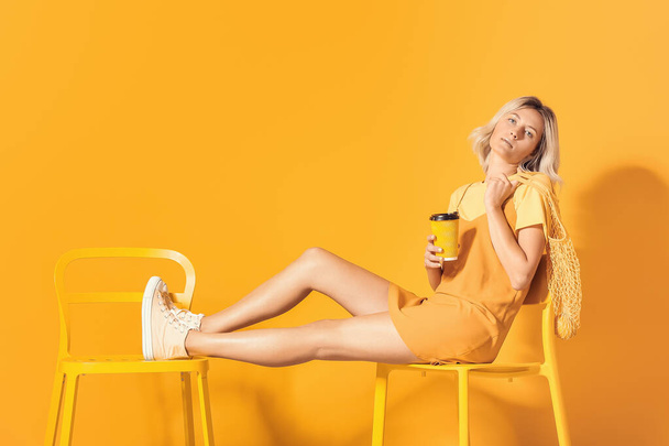Fashionable young woman with coffee sitting on chair near color wall - Photo, image