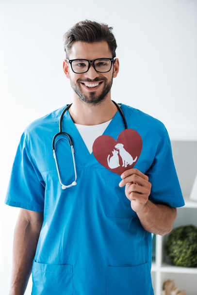 handsome veterinarian smiling at camera while presenting paper cut heart with dog and cat symbols - 写真・画像