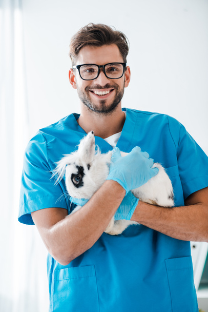schöner Tierarzt lächelt in die Kamera, während er niedliche schwarz-weiße Kaninchen auf Händen hält - Foto, Bild
