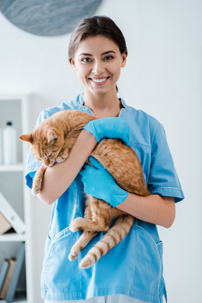 joven veterinario alegre sonriendo a la cámara mientras sostiene lindo gato rojo en las manos
 - Foto, Imagen
