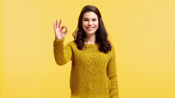 χαμογελαστό κορίτσι σε πουλόβερ δείχνει ok σημάδι απομονώνονται σε κίτρινο - Πλάνα, βίντεο
