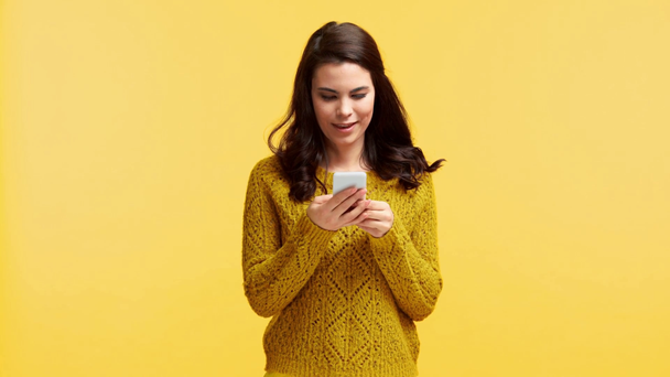 chica sonriente en suéter mensajes de texto en el teléfono inteligente aislado en amarillo
 - Metraje, vídeo