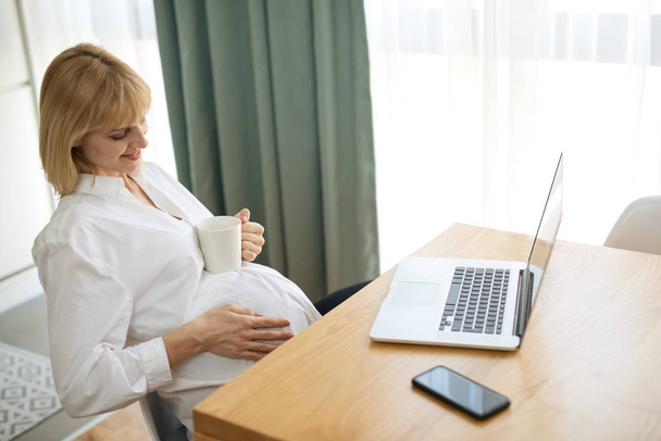 Όμορφη έγκυος επιχειρηματίας και χαμογελώντας, ενώ κάθεται στο χώρο εργασίας στο σπίτι και να πίνουν καφέ - Φωτογραφία, εικόνα