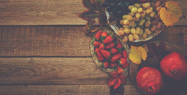 Fresh pomegranates, grapes and berries in an old vintage plate with leaves. On rustic wooden background. Free space for text . Top view. Toned image. - Fotoğraf, Görsel