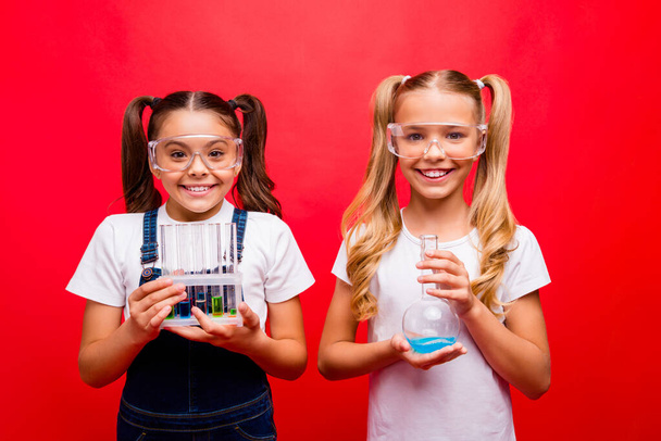 İki küçük kız öğrencinin fotoğrafı kimyasal deney yapıyor. Öğretmenlere tüplerle güvenlik gözlüğü giydiriliyor. Tüm tişörtlerde izole edilmiş kırmızı arka plan rengi var. - Fotoğraf, Görsel
