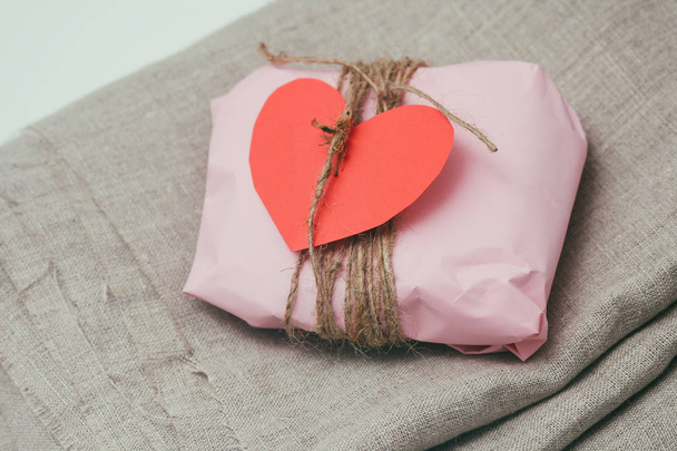 Gift box wrapped in pink craft paper and tie brown string. Carton heart card. Christmas mood. Crumpled brown background. Present package. Delivery service. Soft craft pouch.  - Zdjęcie, obraz