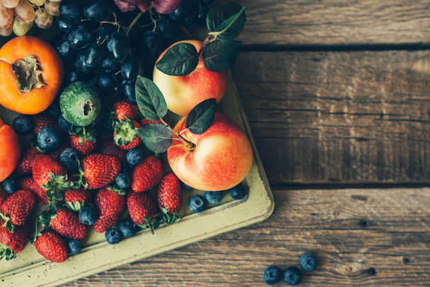 Healthy food clean eating selection in wooden tray: strawberries, blueberries apples, grapes, persimmon, pomegranate on rustic wooden background. Top view. Copy space. - 写真・画像