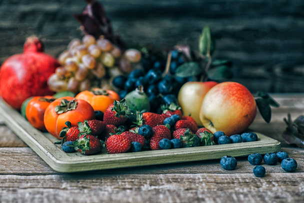 Healthy fruits. Vintage tray filled with strawberries, blueberries apples, grapes, persimmon, pomegranate on rustic wooden background. Toned image. Selective focus. - 写真・画像